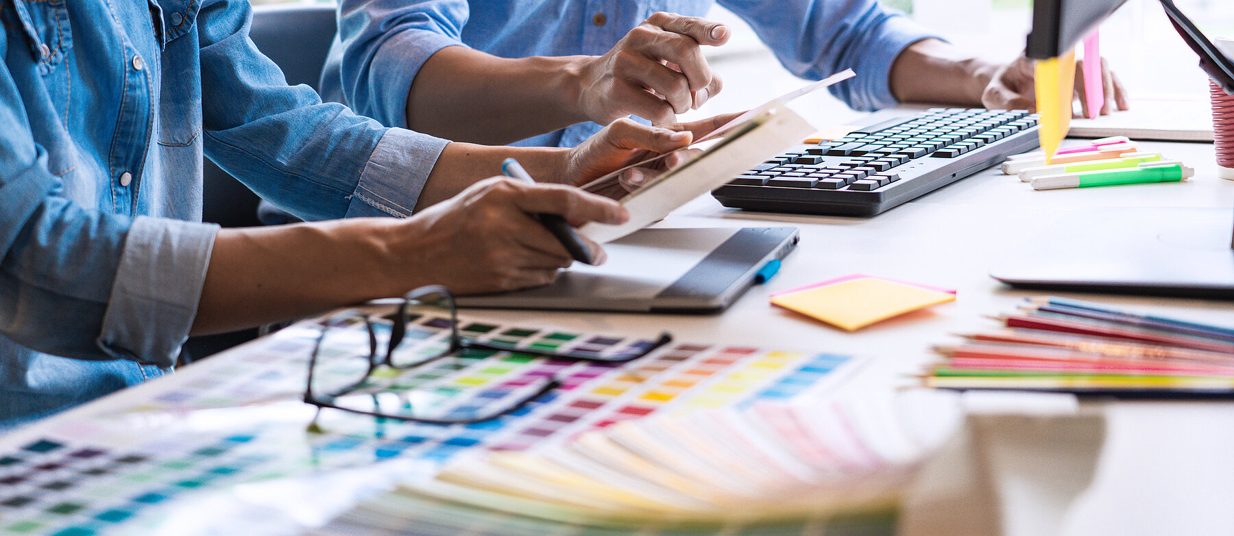 Designer oder Grafiker arbeiten am Schreibtisch mit Farbfächern, Tastatur und Computer – Symbolbild für Grafikdesign oder Farbgestaltung.