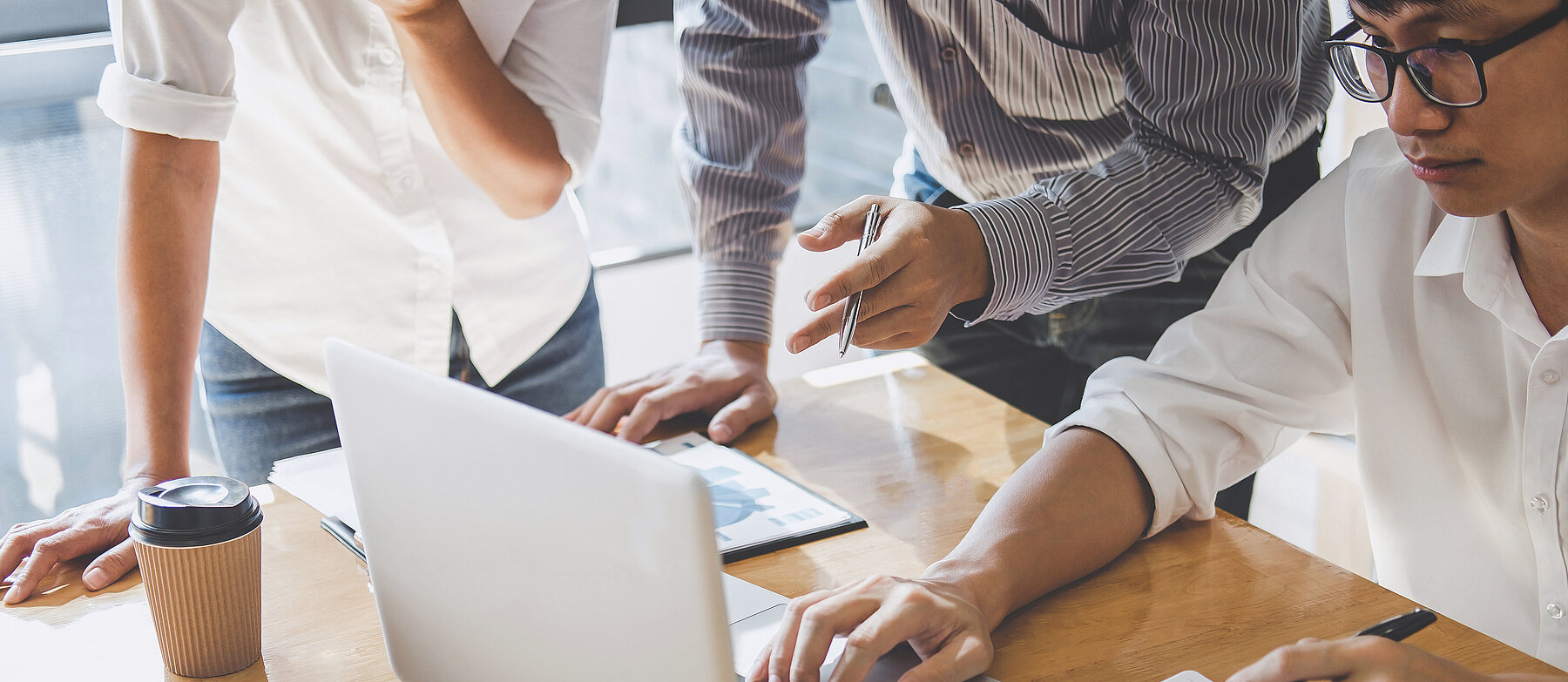 3 Personen arbeiten gemeinsam an einem Holztisch mit Laptop, Buch und Tablet; eine Person tippt, während die anderen etwas zeigen und besprechen.
