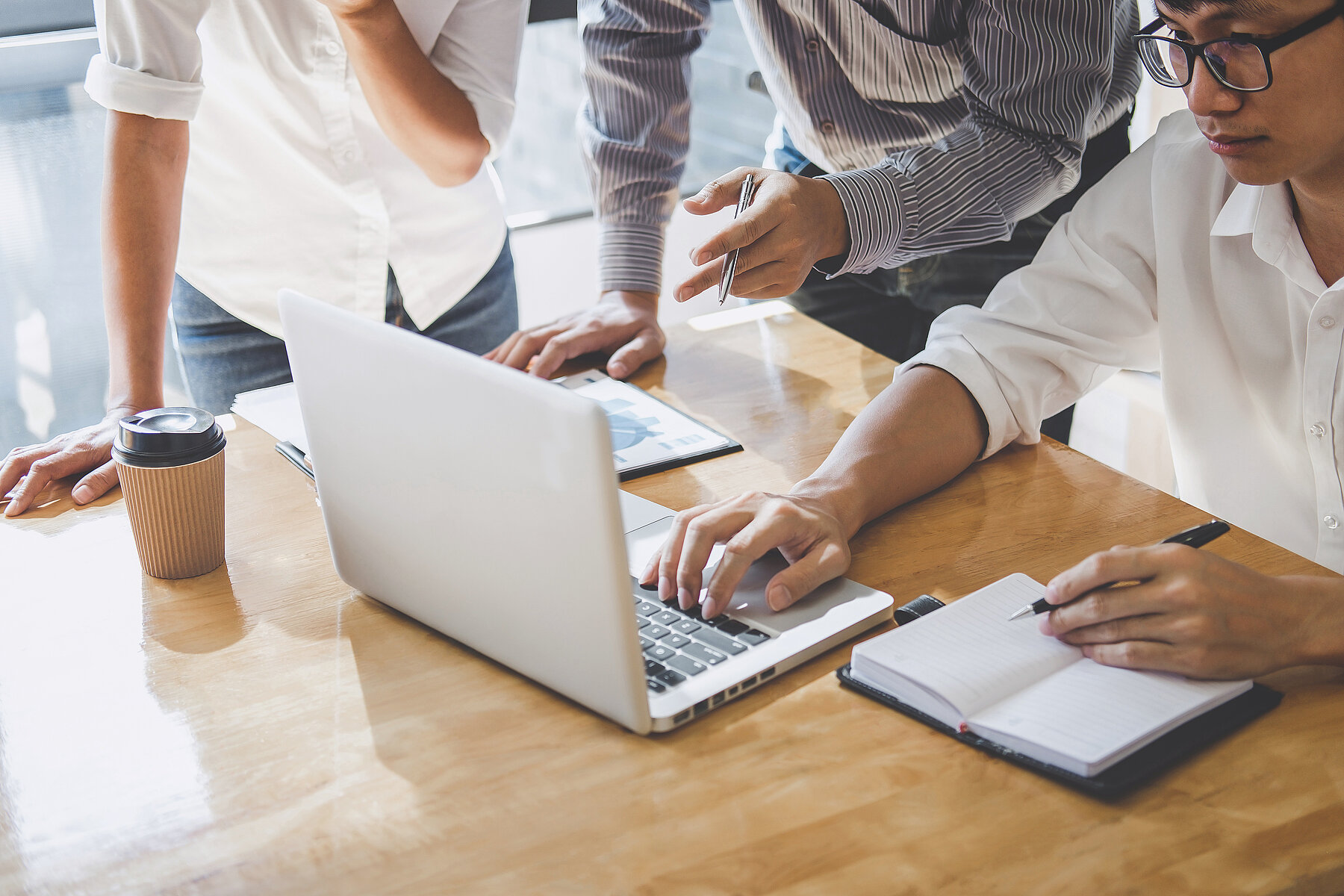 3 Personen arbeiten gemeinsam an einem Tisch mit Laptop, Block und Kaffeebecher; eine Person tippt, während die anderen auf den Bildschirm zeigen.
