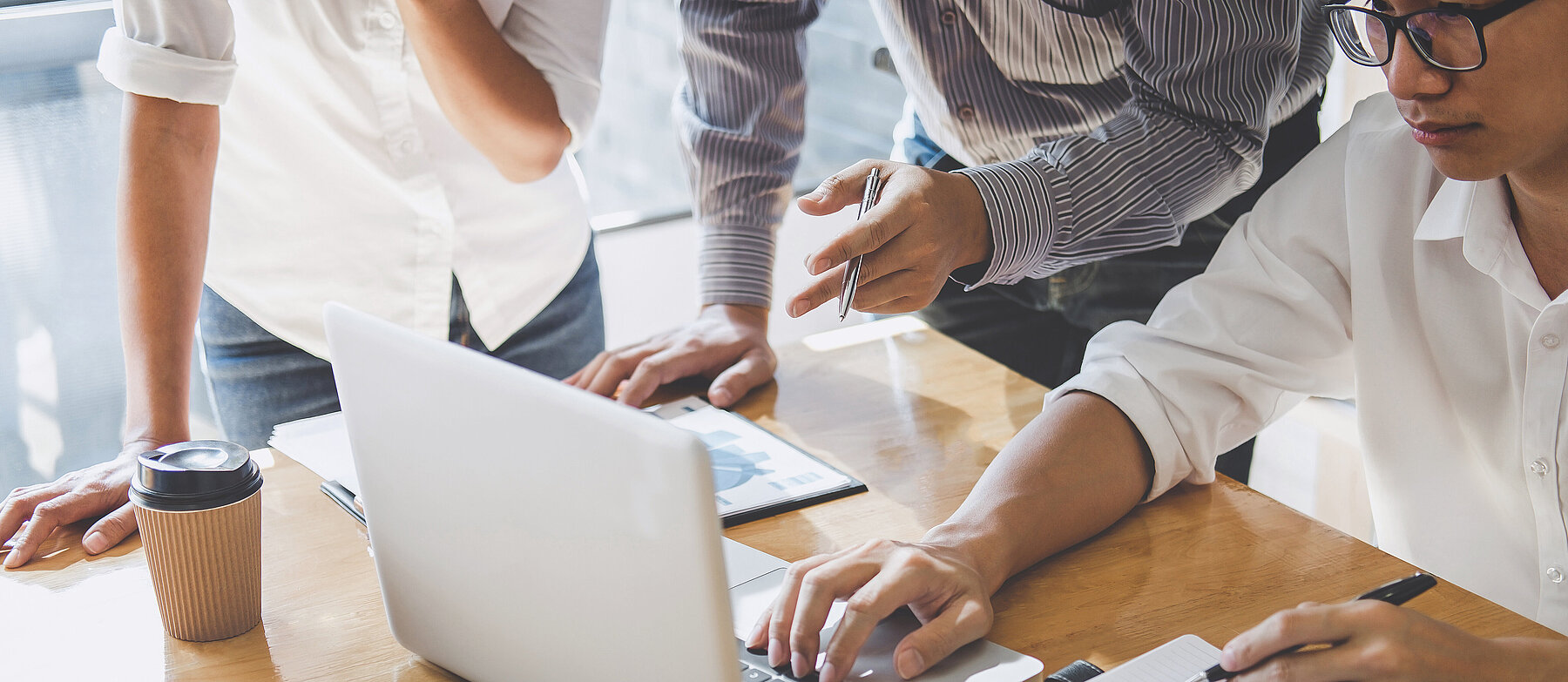 3 Personen arbeiten gemeinsam an einem Holztisch mit Laptop, Buch und Tablet; eine Person tippt, während die anderen etwas zeigen und besprechen.