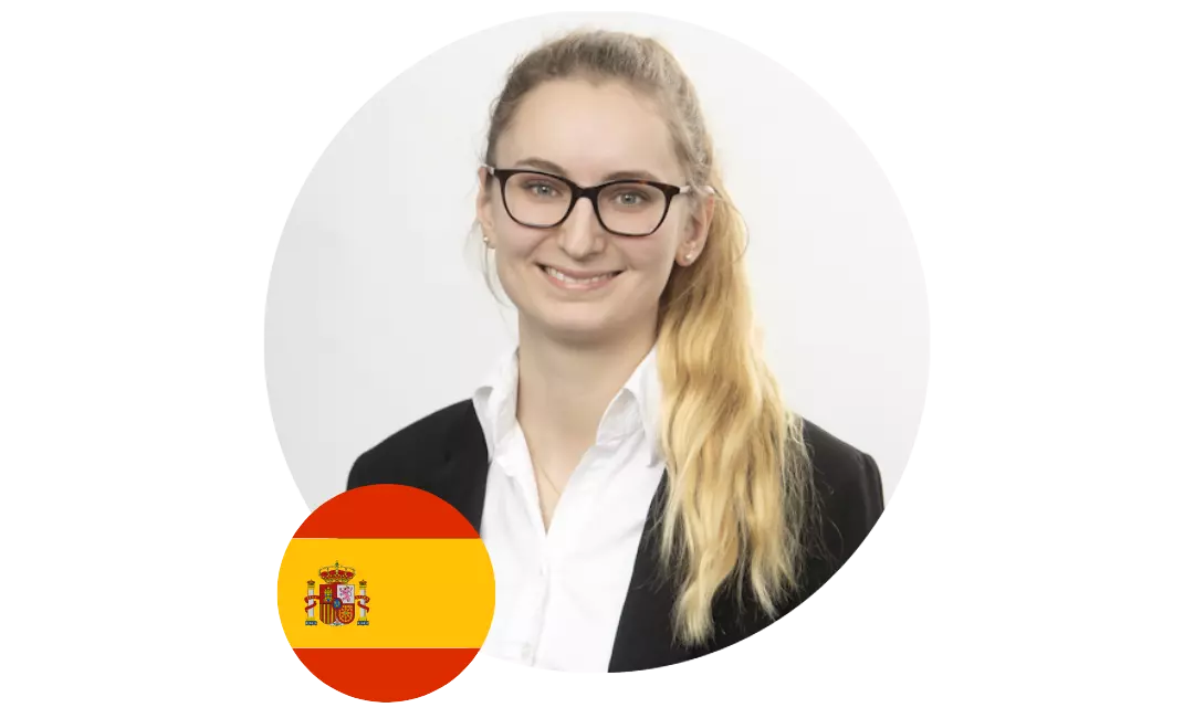 Porträt einer lächelnden jungen Frau mit Brille vor hellem Hintergrund, im unteren Bildbereich eine spanische Flagge.