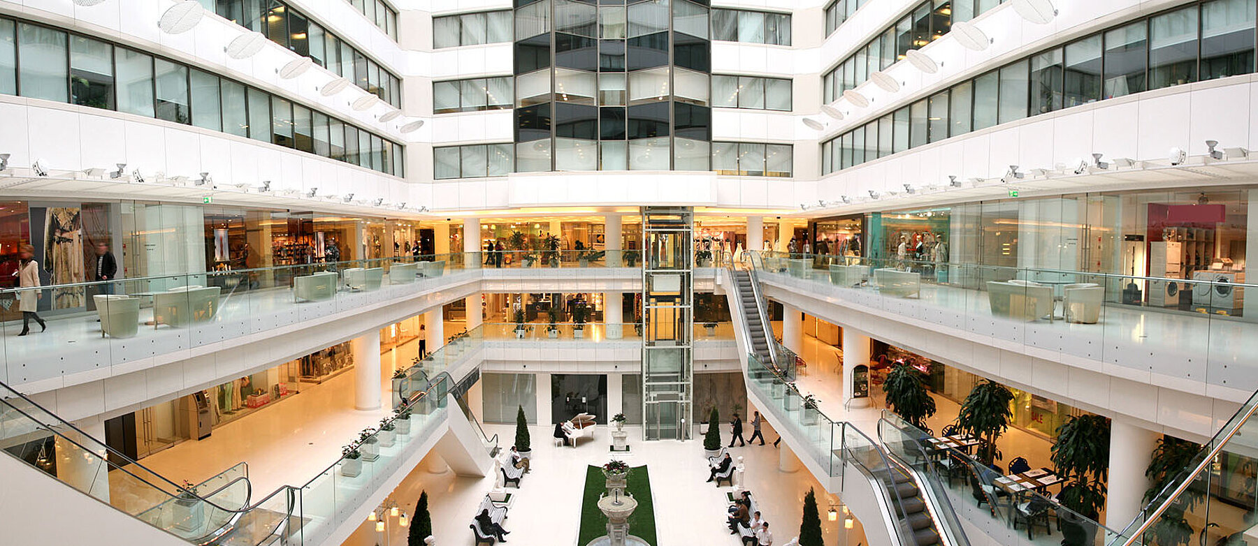 Mehrstöckiges Einkaufszentrum mit gläsernen Geländern, Rolltreppen und offenen Ladenflächen in heller, lichtdurchfluteter Architektur.
