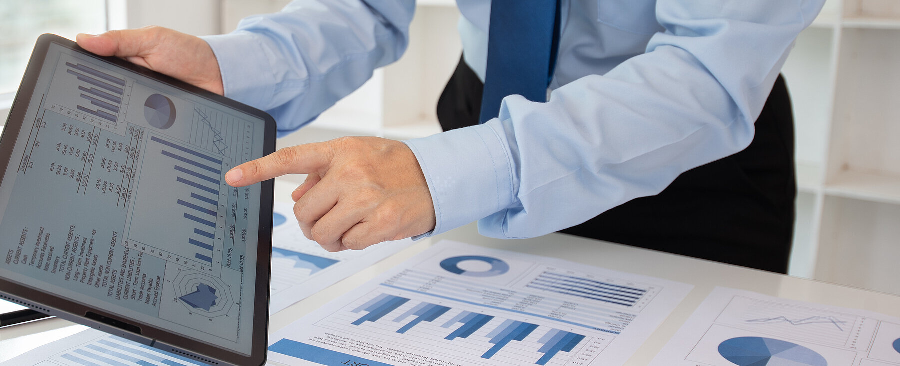 Geschäftsperson zeigt auf Diagramme; auf dem Tisch liegen weitere Finanzberichte und ein Laptop– Symbolbild für Datenanalyse.