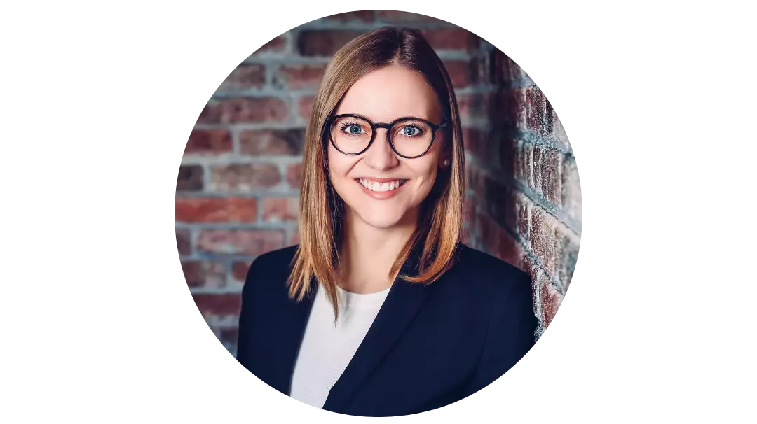 Geschäftsfrau mit braunen Haaren die vor einer Ziegelsteinwand in schwarz-weißem Anzug und Brille lächelnd in die Kamera schaut.