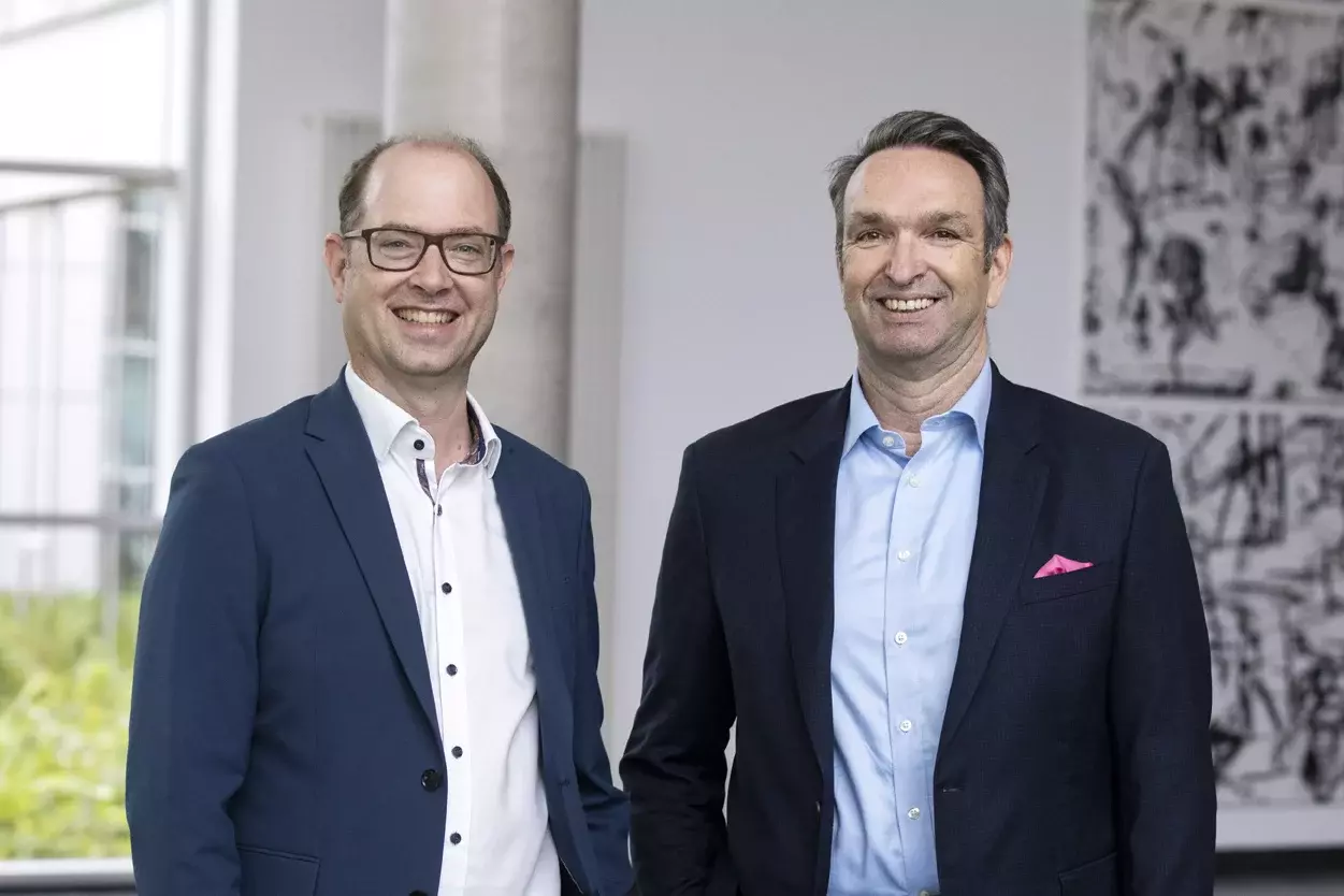 Zwei Männer im Business-Outfit stehen lächelnd nebeneinander in einem modernen Büro.