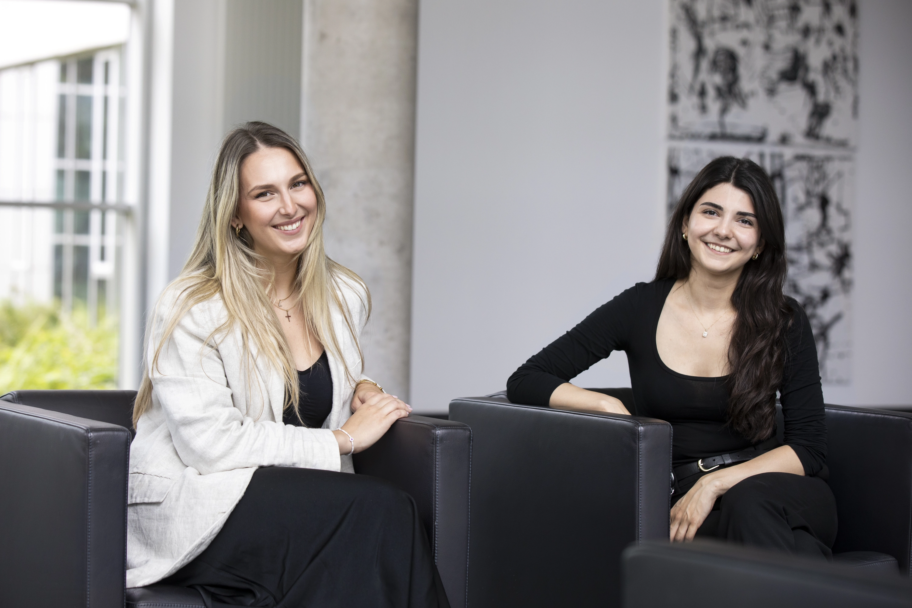 Zwei junge Frauen sitzen lächelnd in schwarzen Sesseln in einem modernen, hellen Büro oder Empfangsbereich.