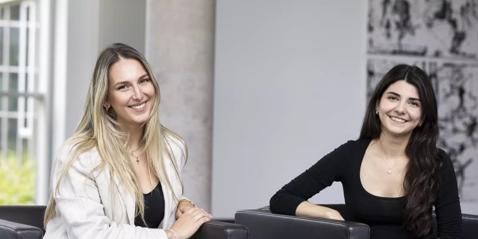Zwei junge Frauen sitzen lächelnd in schwarzen Sesseln in einem modernen, hellen Büro oder Empfangsbereich.