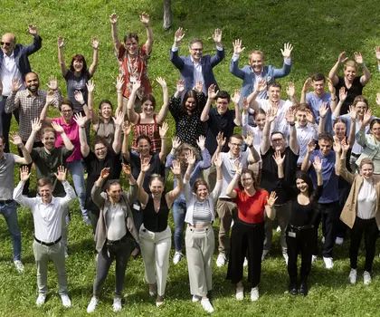 Große Gruppe von Menschen steht auf einer Wiese und winkt fröhlich in die Kamera – Gemeinschaftsfoto bei sonnigem Wetter.