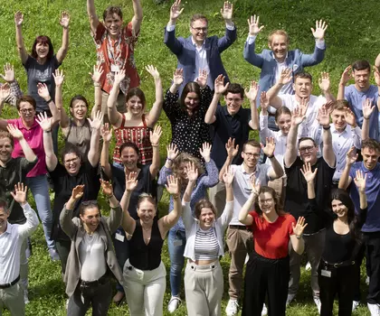 Gruppenfoto von Mitarbeitenden im Freien, die fröhlich in die Kamera winken.