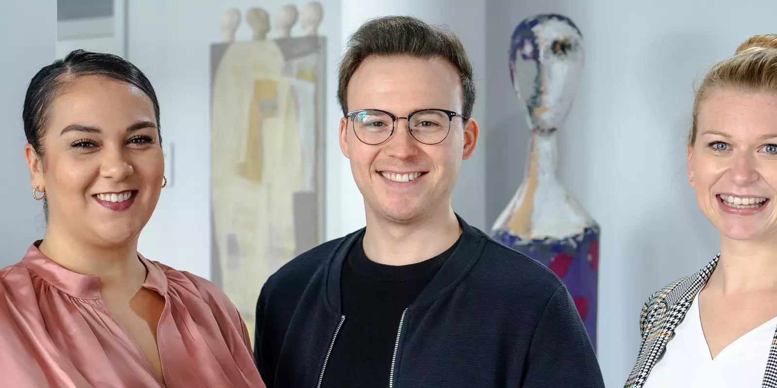 3 Personen stehen lächelnd nebeneinander in einem hellen Flur mit medizinischen Postern – Symbol für Team in einer medizinischen Einrichtung.