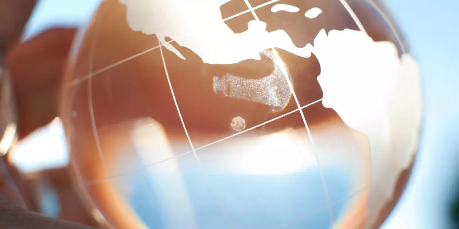 Hand hält eine transparente Glaskugel mit Weltkartenmotiv, im Hintergrund blauer Himmel und Bäume.