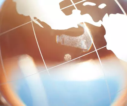 Hand hält eine transparente Glaskugel mit Weltkartenmotiv, im Hintergrund blauer Himmel und Bäume.