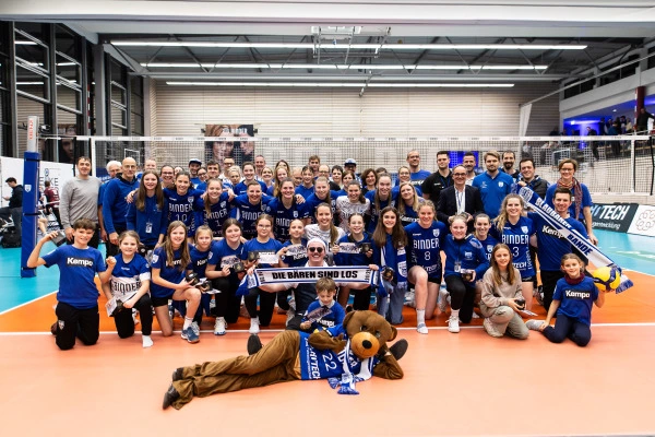 Großes Gruppenfoto eines Volleyballteams mit Trainerstab in einer Sporthalle, alle tragen blaue Trikots und posieren gemeinsam vor dem Netz.