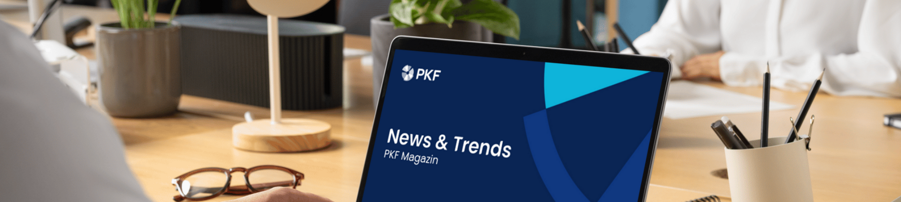 Laptop auf einem Schreibtisch zeigt eine Präsentation mit dem Titel „News & Trends“ der DATEV, im Hintergrund Personen in einem modernen Büro.