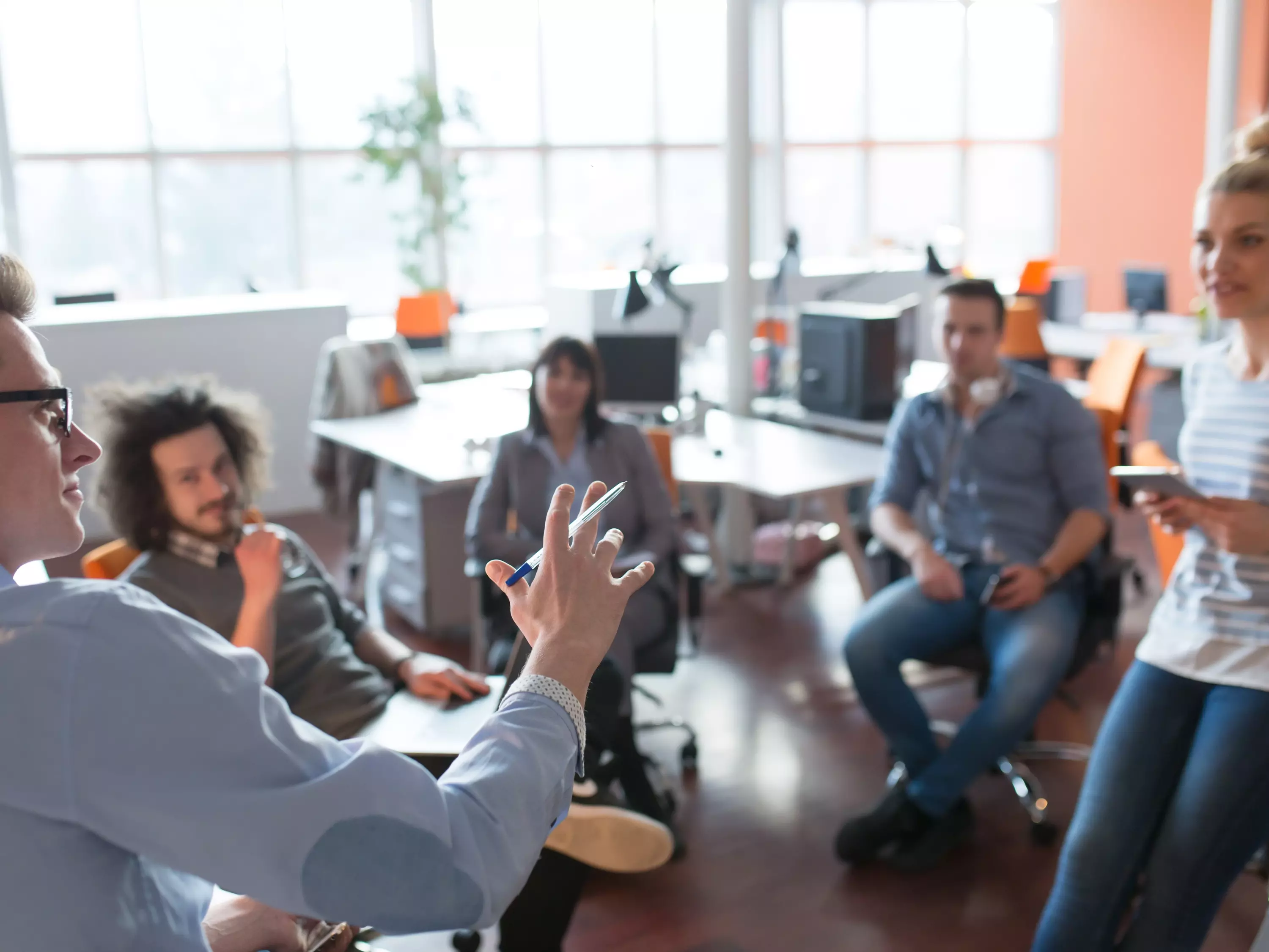 Gruppe junger Menschen in einem Büro sitzt im Kreis und hört einer Person aufmerksam zu, die spricht – Symbol für Teammeeting oder Brainstorming.