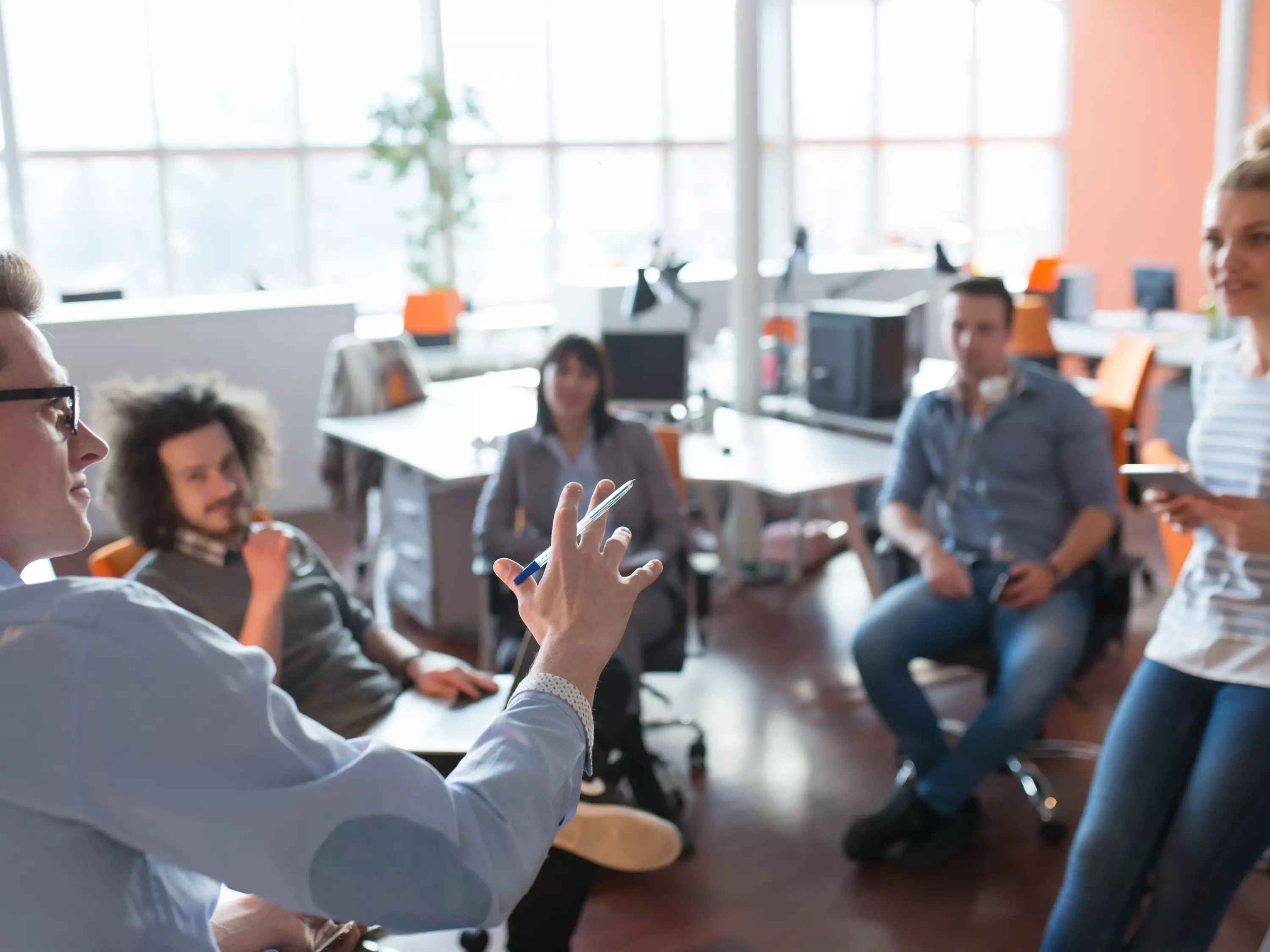 Gruppe junger Menschen in einem Büro sitzt im Kreis und hört einer Person aufmerksam zu, die spricht – Symbol für Teammeeting oder Brainstorming.