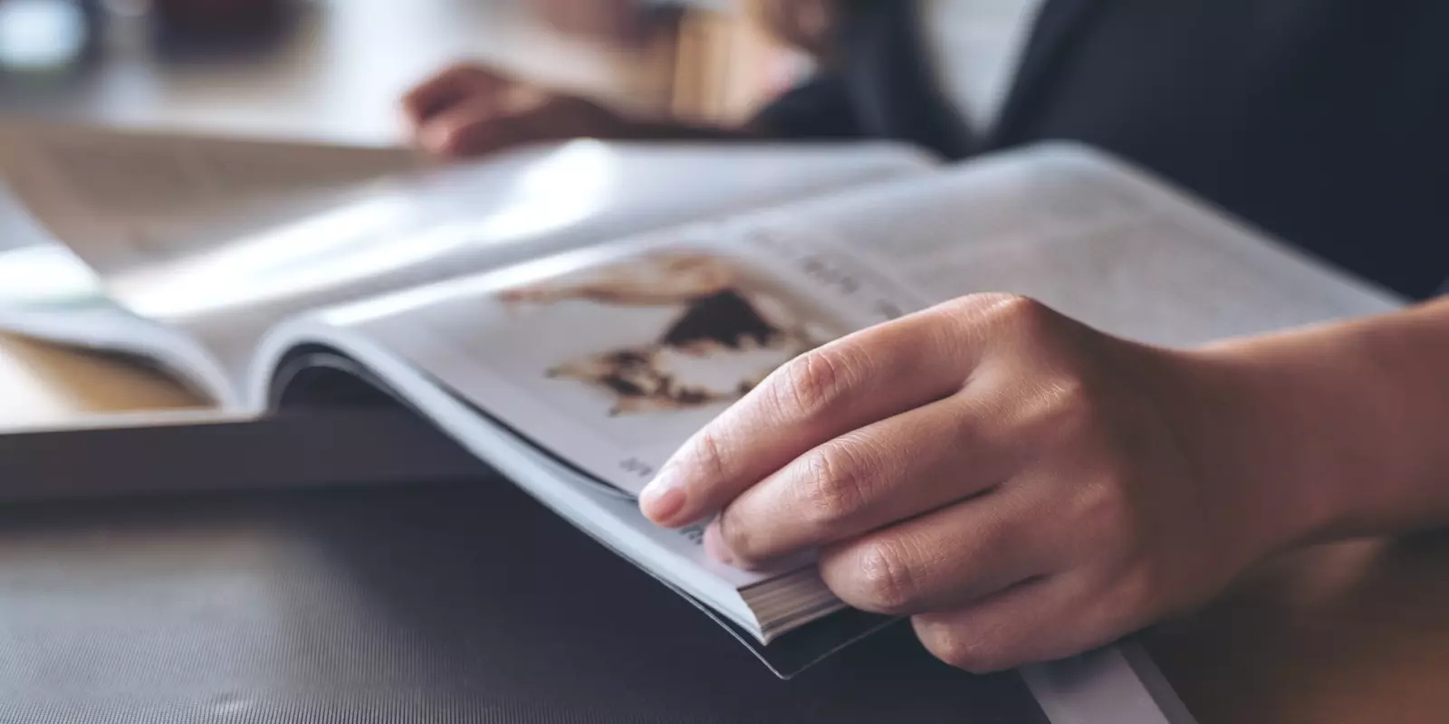 Person blättert in einer Zeitschrift an einem Tisch mit Laptop und Getränk im Hintergrund.