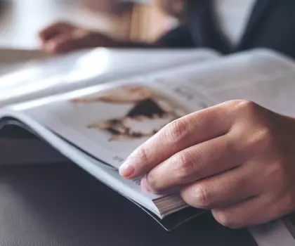 Person blättert in einer Zeitschrift an einem Tisch mit Laptop und Getränk im Hintergrund.