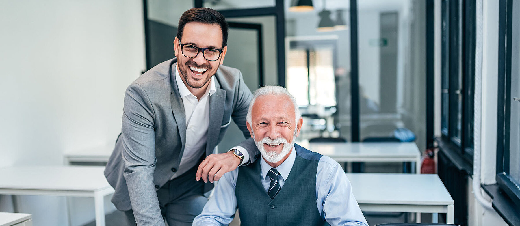 2 Geschäftsleute unterschiedlichen Alters in einem Büro; einer sitzt am Tisch, der andere steht daneben und legt ihm die Hand auf die Schulter.