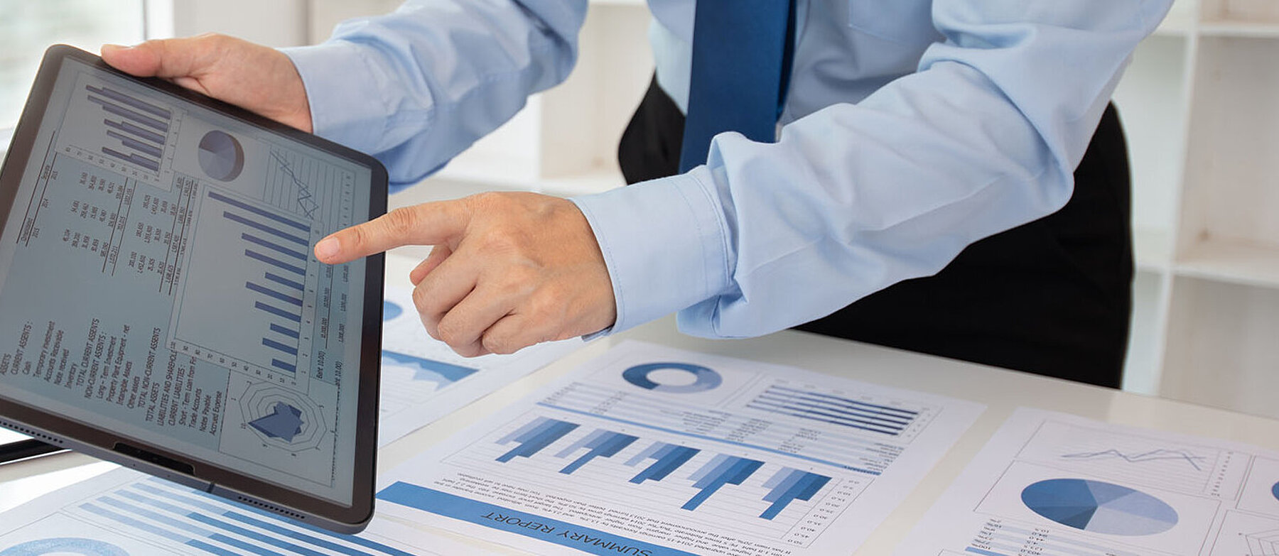 Geschäftsperson zeigt auf ein Tablet mit digitalen Diagrammen; auf dem Tisch liegen weitere Geschäftsgrafiken und Datenanalysen in Blau.