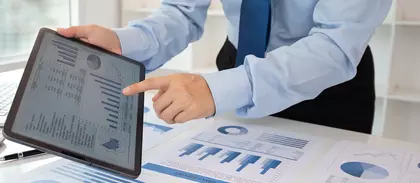 Geschäftsperson im Hemd und Krawatte zeigt auf ein Tablet mit Diagrammen und Statistiken; auf dem Tisch liegen gedruckte Auswertungen in Blau.