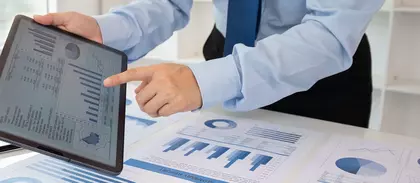 Geschäftsperson zeigt auf ein Tablet mit digitalen Diagrammen; auf dem Tisch liegen weitere Geschäftsgrafiken und Datenanalysen in Blau.