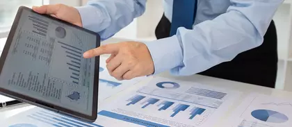 Geschäftsperson zeigt auf ein Tablet mit digitalen Diagrammen; auf dem Tisch liegen weitere Geschäftsgrafiken und Datenanalysen in Blau.