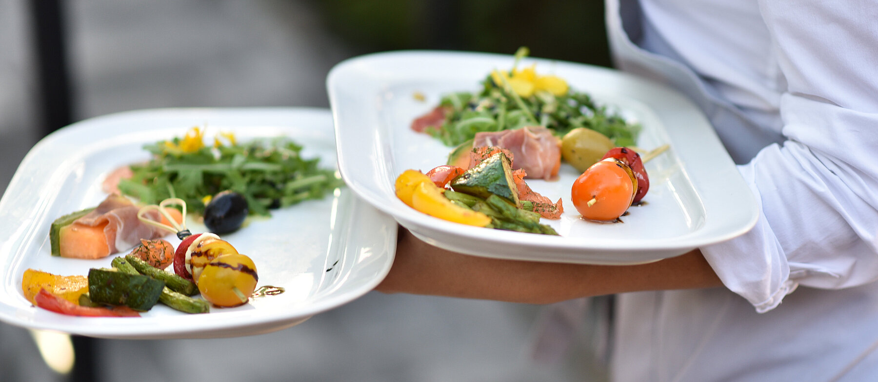 Kellner:in serviert zwei weiße Teller mit bunt angerichteten, hochwertigen Speisen – Symbolbild für Gastronomie oder gehobene Küche.