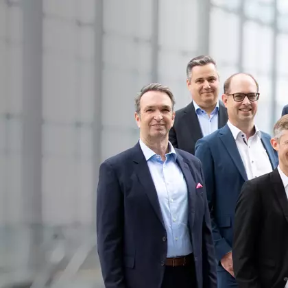 Geschäftsleute stehen vor einer Glasfassade und blicken lächelnd in die Kamera – Symbolbild für Führungsteam.