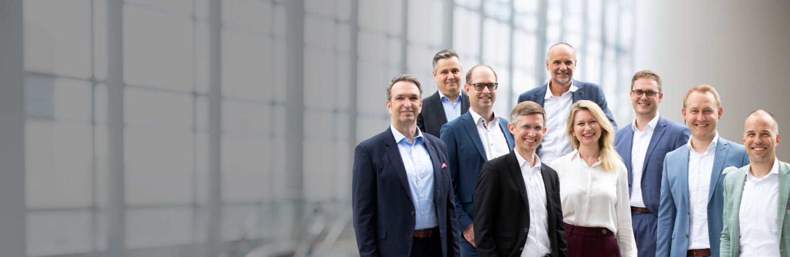 Geschäftsleute stehen vor einer modernen Glasfassade und blicken lächelnd in die Kamera – Symbolbild für Führungsteam oder Unternehmensgruppe.