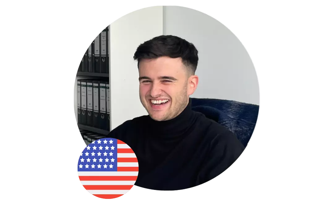 Lächelnder junger Mann in dunklem Rollkragenpullover vor einem hellen Hintergrund, unten links im Bild ist eine US-Flagge eingeblendet.