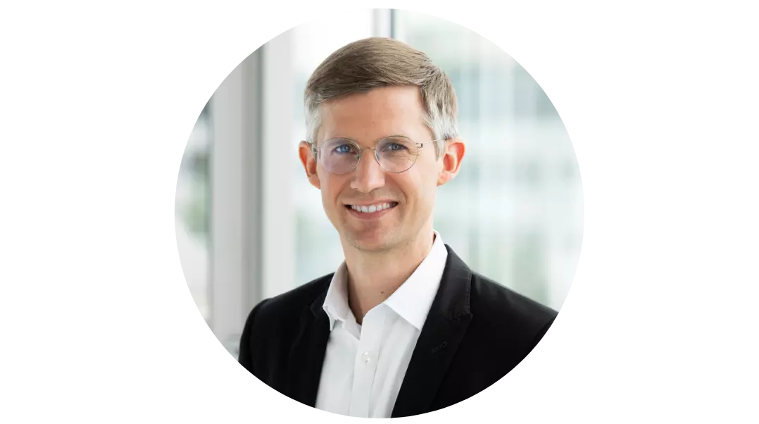 Geschäftsmann in schwarz-weißem Anzug der freundlich lächelnd in die Kamera schaut.