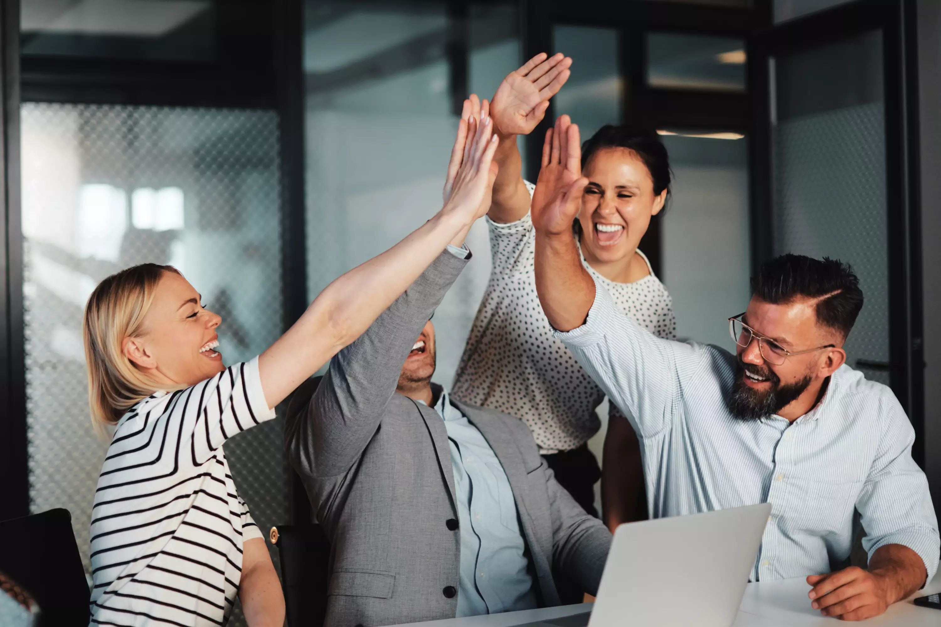 4 junge Berufstätige in einem modernen Büro feiern gemeinsam mit einem High-Five – Symbolbild für Teamwork, Erfolg und Zusammenarbeit.
