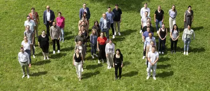 Gruppe von Menschen steht in lockerer Formation auf einer Wiese und blickt zur Kamera – Symbol für Team, Gemeinschaft oder Mitarbeitende im Freien.