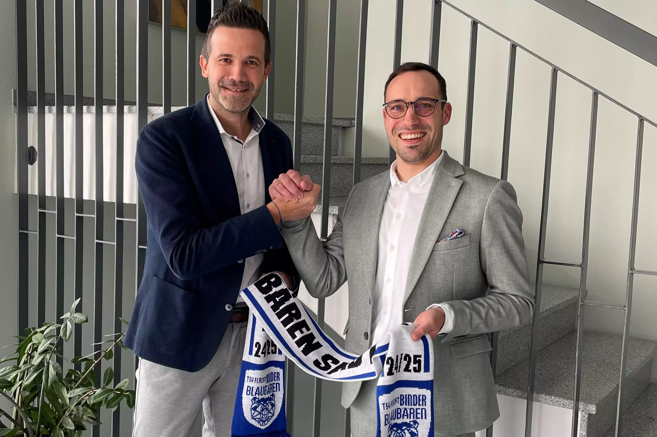 2 Männer im Business-Outfit stehen lächelnd nebeneinander und halten ein blau-weißes Volleyballtrikot – symbolisch für eine Partnerschaft.