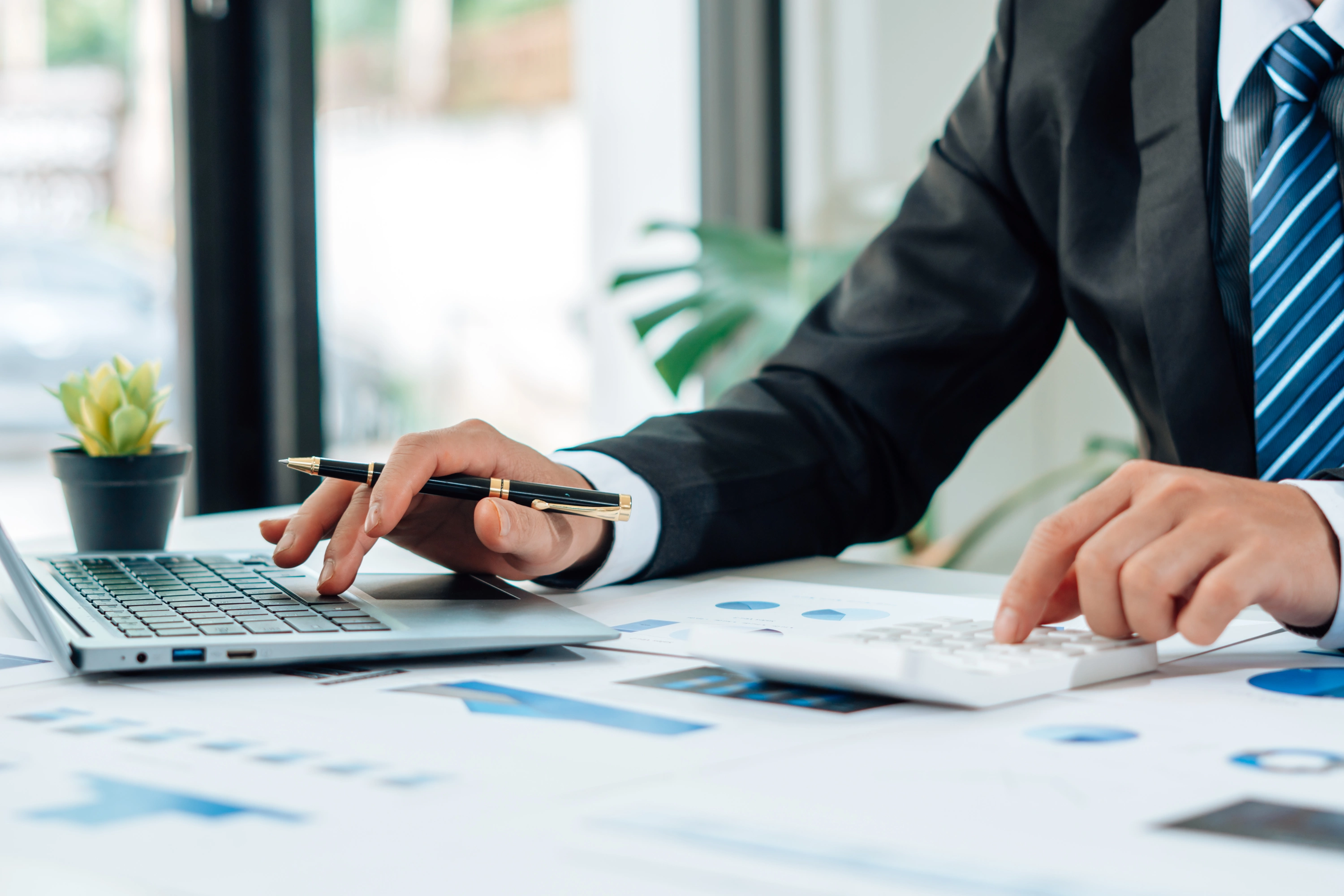 Geschäftsperson arbeitet am Schreibtisch mit Laptop, Taschenrechner und Finanzunterlagen- Symbol für Buchhaltung oder Finanzanalyse.