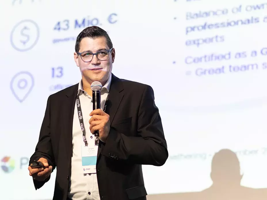 Mann in Businesskleidung hält einen Vortrag mit Mikrofon vor einer Projektionswand mit Diagrammen und Text.