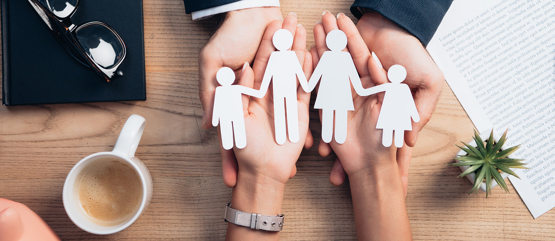 2 Hände halten symbolisch ausgeschnittene Papierfiguren einer Familie über einem Schreibtisch– Symbolbild für Versicherungsschutz.