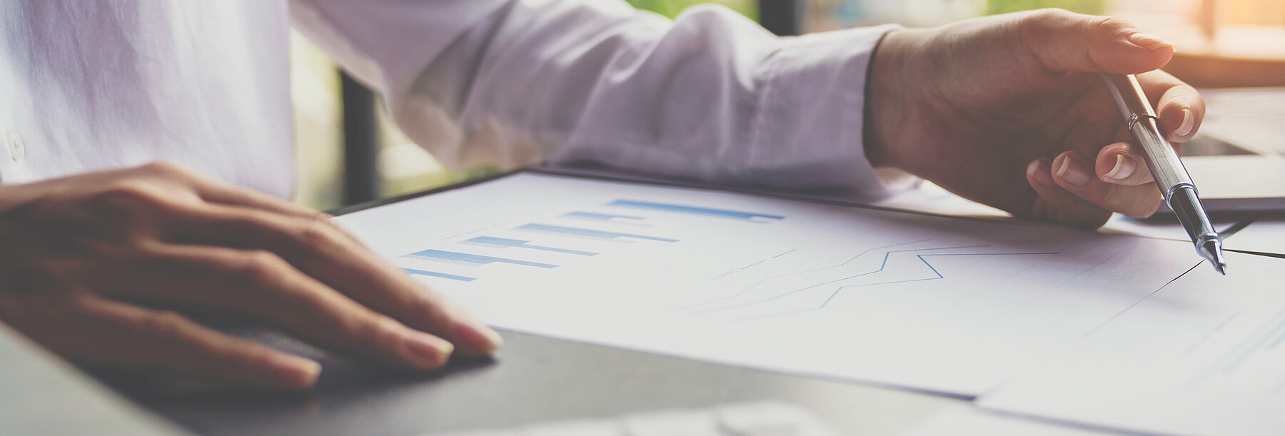 Person analysiert ein Dokument mit Diagrammen an einem Schreibtisch und zeigt mit einem Stift auf bestimmte Daten – Symbol für Finanzanalyse.