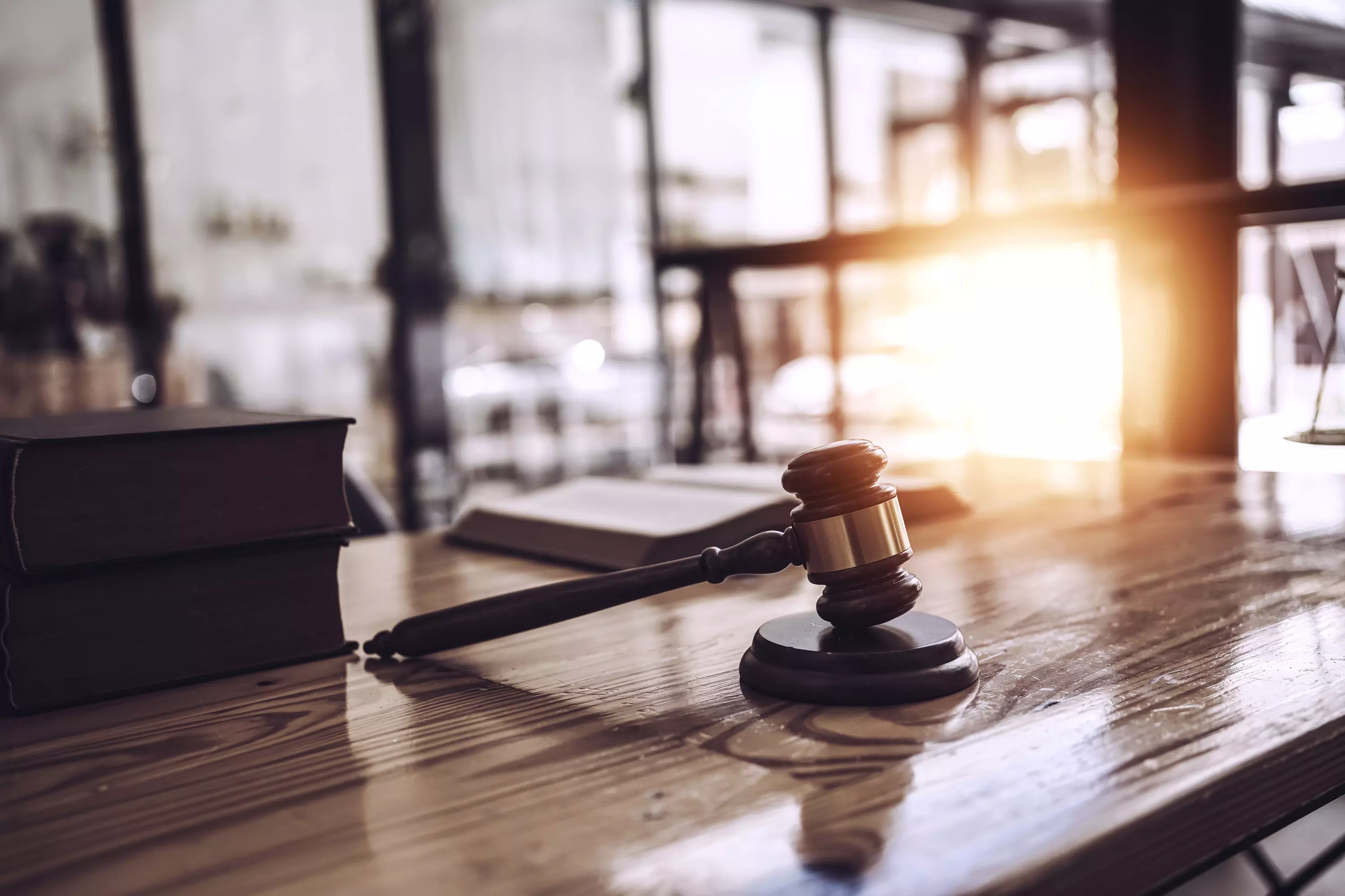 Modernes Büro oder Konferenzraum mit Glaswänden und großem Holztisch, auf dem ein Richterhammer liegt; Sonnenlicht strahlt durch das Fenster.