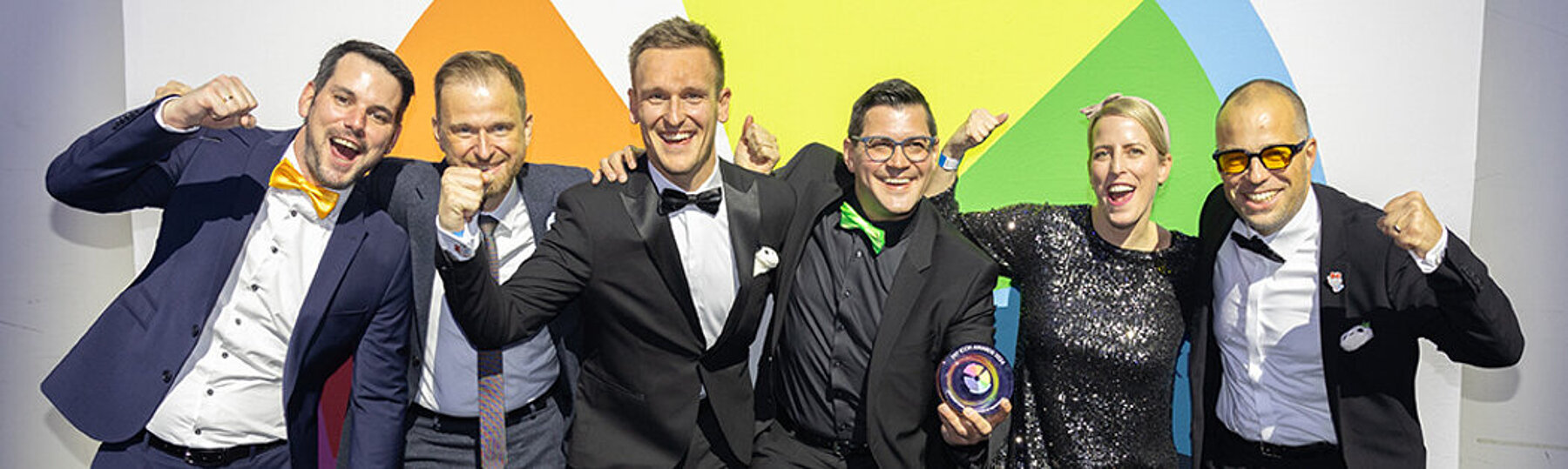 Gruppe elegant gekleideter Menschen posiert fröhlich vor einer bunten Wand bei einer Preisverleihung oder Gala.