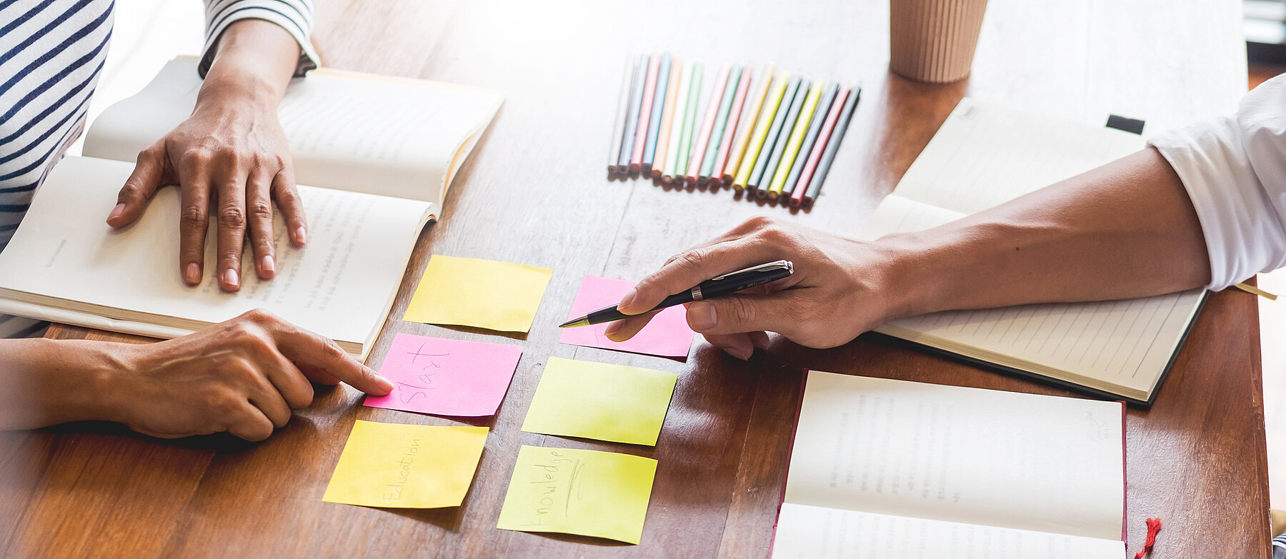2 Personen arbeiten gemeinsam an einem Tisch mit Büchern und bunten Klebezetteln – Symbolbild für Planung, Organisation oder kreatives Arbeiten.