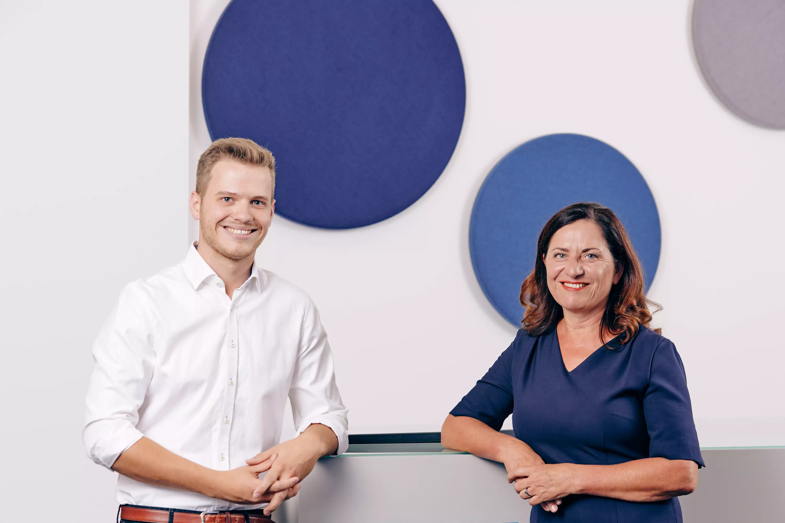 Zwei Personen stehen vor einer Wand mit dekorativen Kreisen und lächeln in die Kamera.