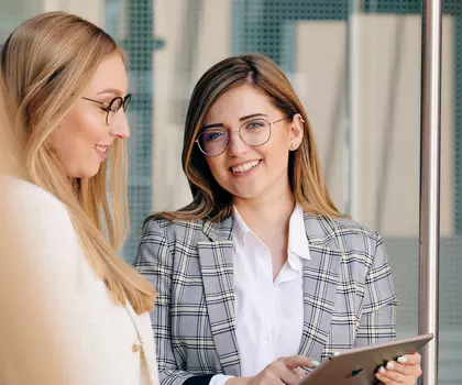 Zwei Geschäftsfrauen im Gespräch in einem Büro, eine von ihnen hält ein Tablet – Symbolbild für Zusammenarbeit oder Beratung im Business-Kontext.