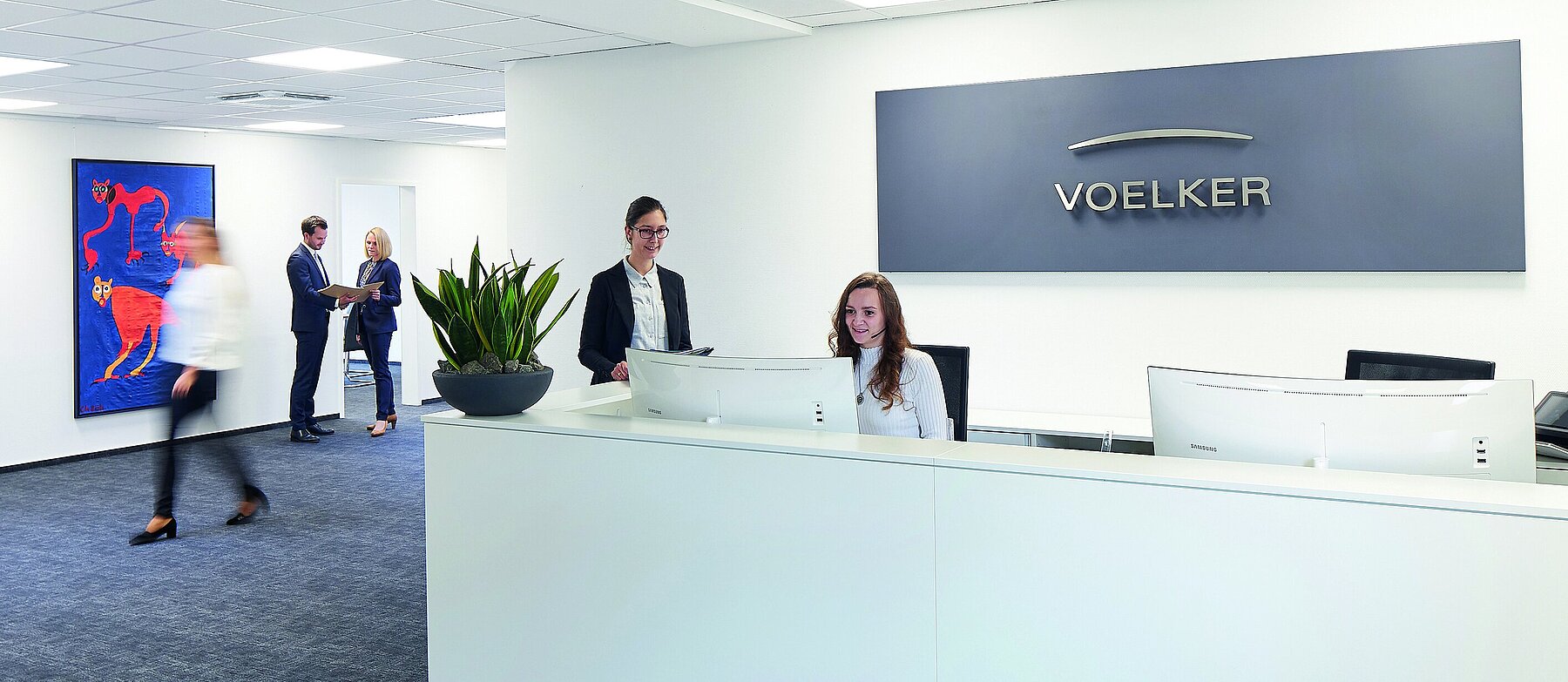 Empfangsbereich eines Büros mit weißem Tresen, 2 Empfangsmitarbeitenden, Firmenlogo an der Wand und mehreren Geschäftsleuten im Hintergrund.