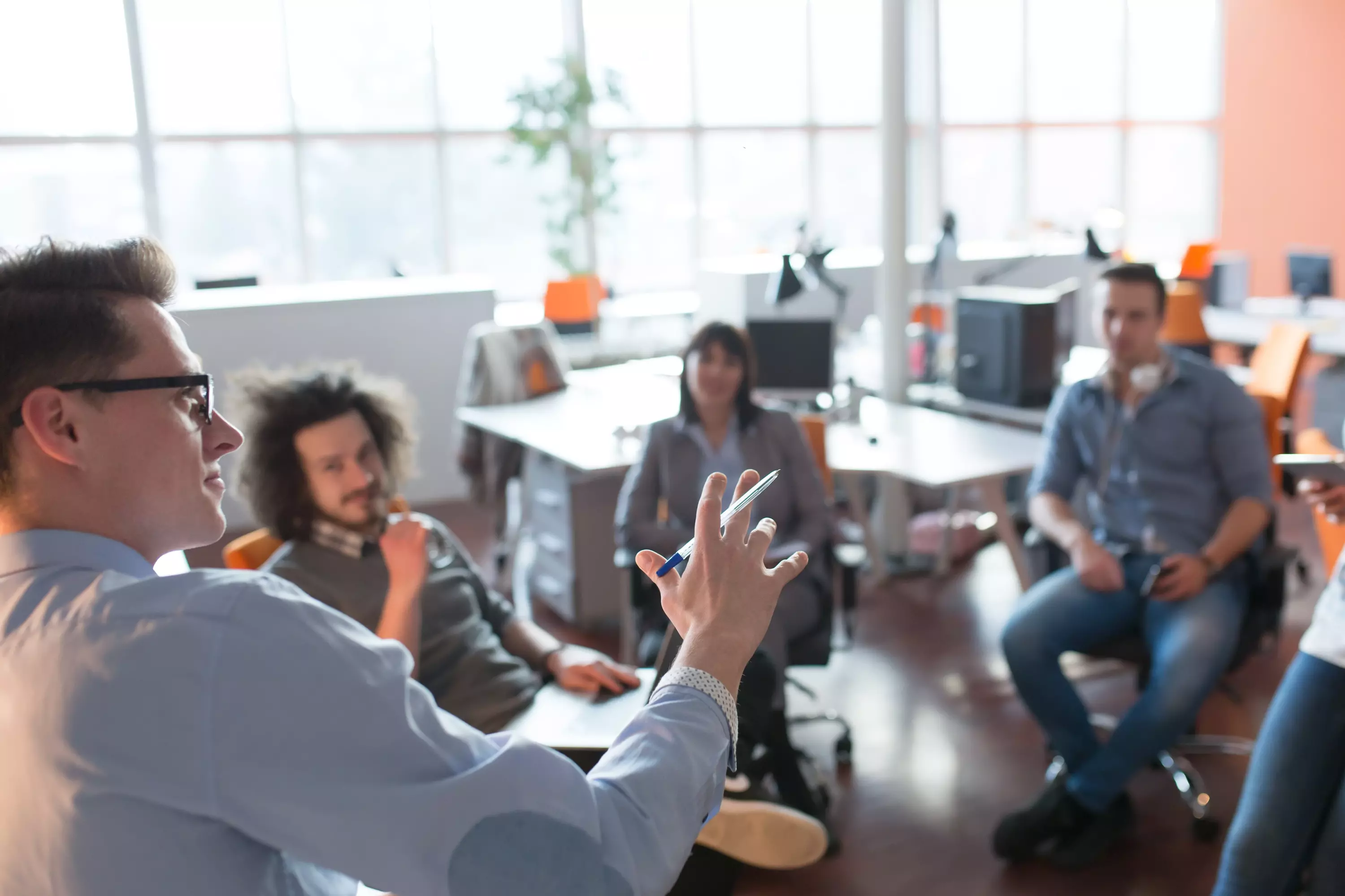 Junge Menschen sitzen in einem Büro in einer Gesprächsrunde, eine Person spricht– Symbolbild für Teammeeting oder kreative Zusammenarbeit.