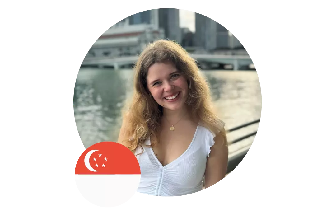 Profilbild einer lächelnden jungen Frau vor einer urbanen Wasserlandschaft, mit einer Singapur-Flagge im Vordergrund.