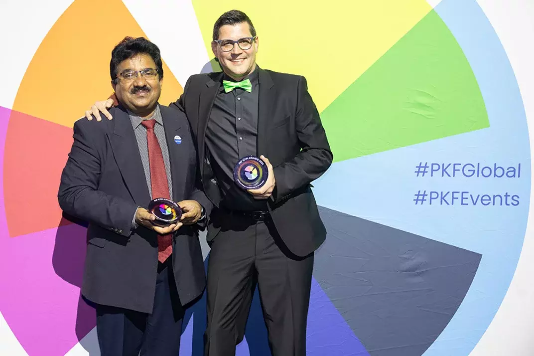 2 Männer in Anzügen posieren mit Auszeichnungen vor einem farbenfrohen Kreisdiagramm-Hintergrund bei einer Preisverleihung oder einem Event.