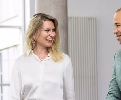 Zwei beruflich gekleidete Personen, eine Frau und ein Mann, unterhalten sich freundlich in einem modernen Bürogebäude.