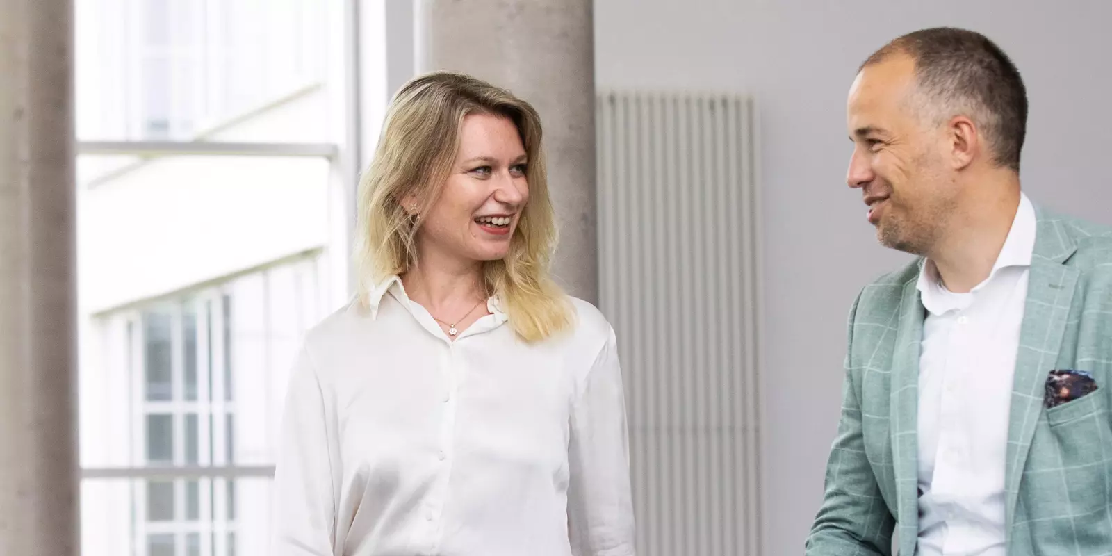 Zwei beruflich gekleidete Personen, eine Frau und ein Mann, unterhalten sich freundlich in einem modernen Bürogebäude.